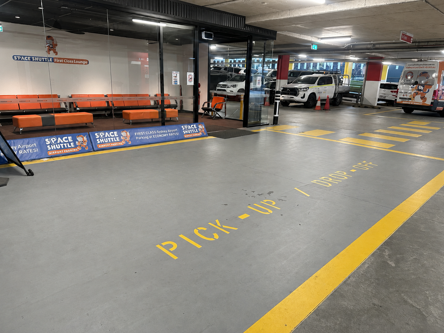 Airport pickup zone with orange seating area behind glass and signs for Space Shuttle airport parking services nearby