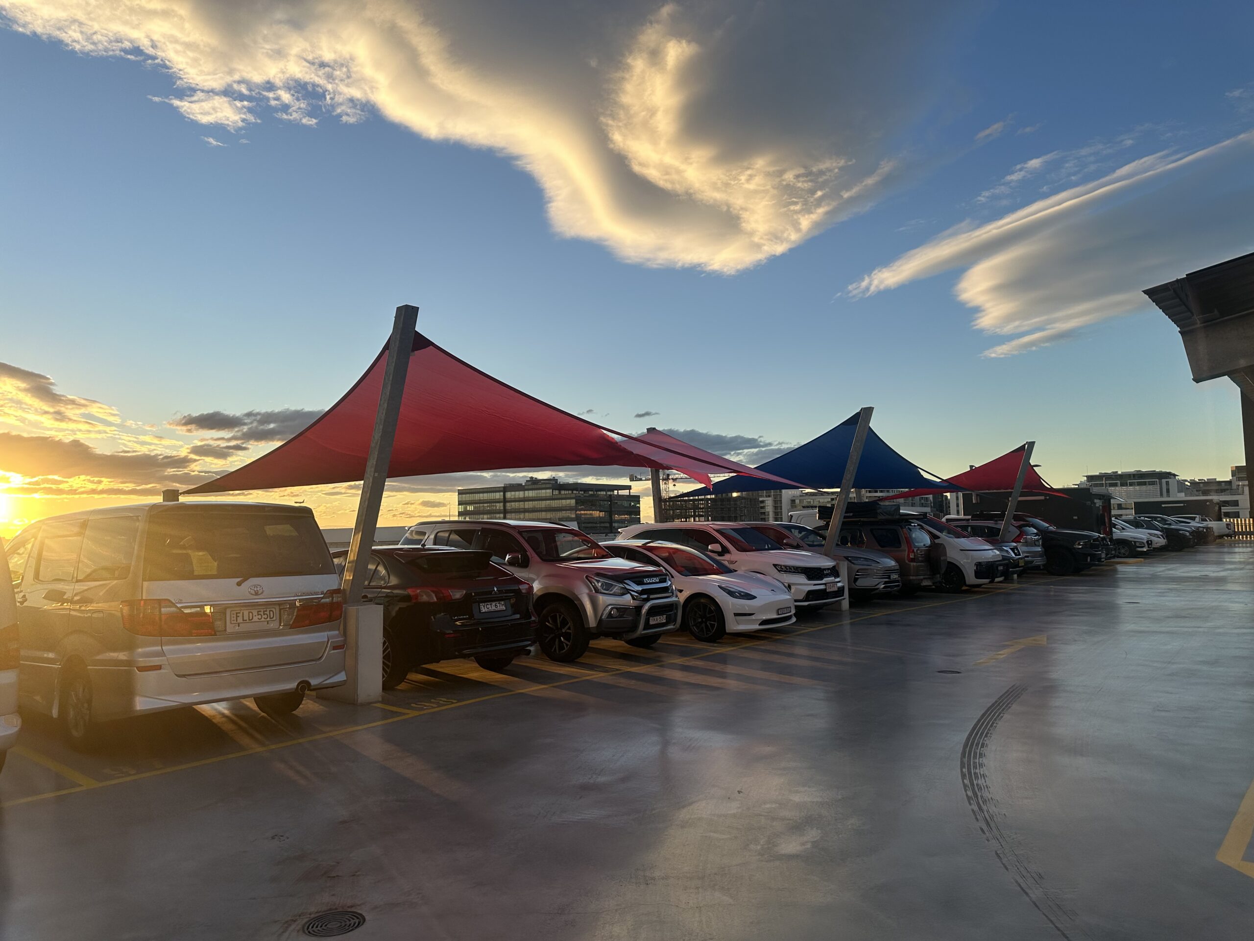 Sunset over a rooftop parking lot with red and blue shade sails and many parked cars.
