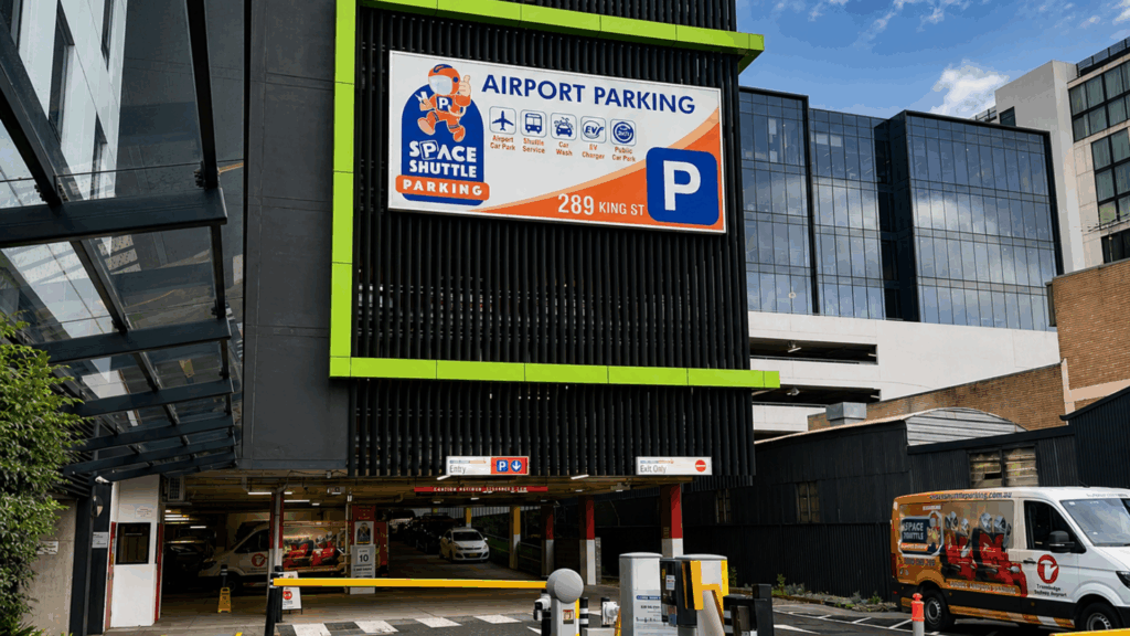 Front view of a parking garage entrance with a large 'Airport Parking' sign, Space Shuttle Parking logo, and a blue P symbol on the sign nearby.