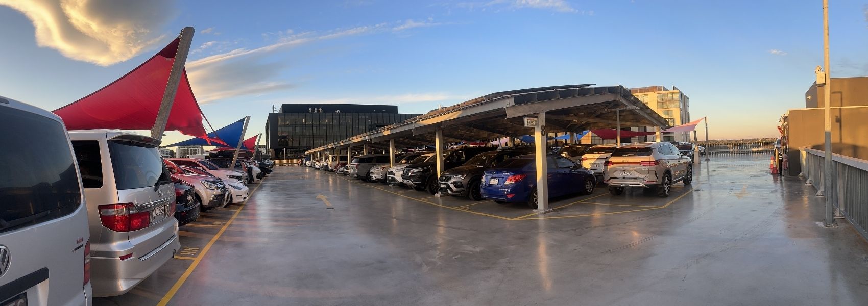 Panoramic view of a rooftop parking deck with cars parked under red and blue shade sails at sunset.