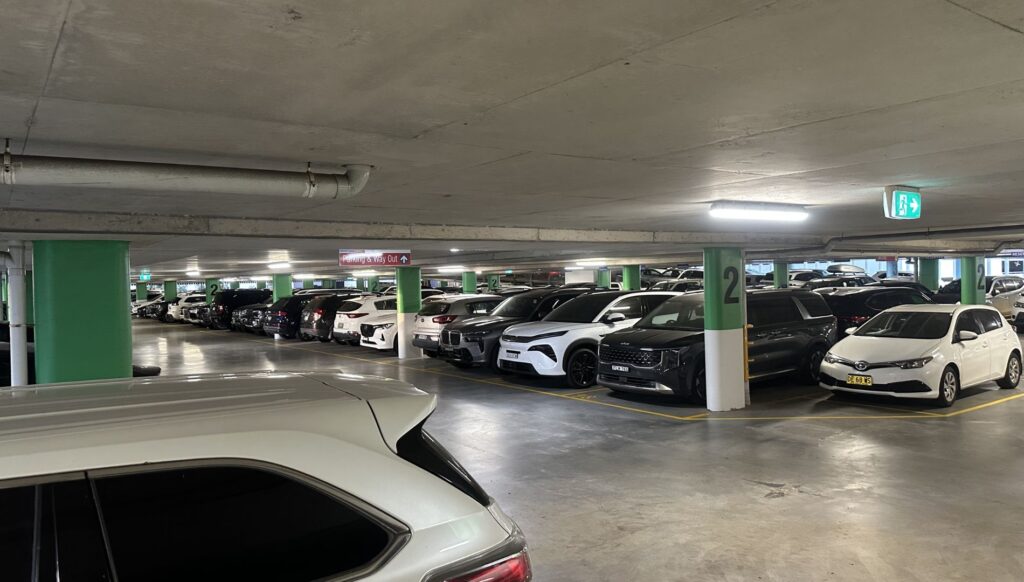 Underground parking garage with rows of cars, green support pillars, and exit signs nearby.
