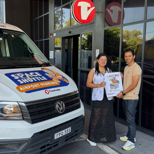 Two people stand outside a glass-front building, smiling while holding an 'I Just Won' prize poster next to a white van with an airport parking sticker, implying a prize win.