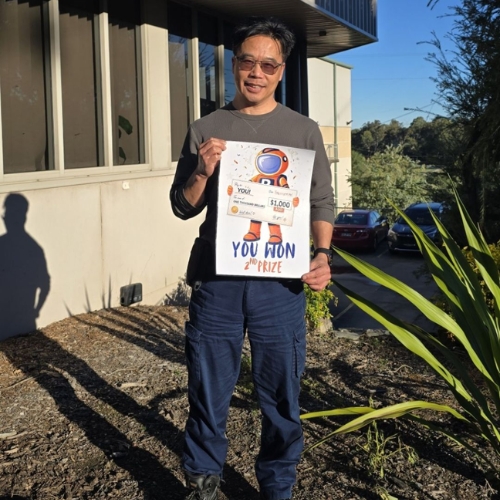 Man wearing sunglasses and a grey shirt outdoors, proudly holding a certificate reading You Won $1,000 prize.