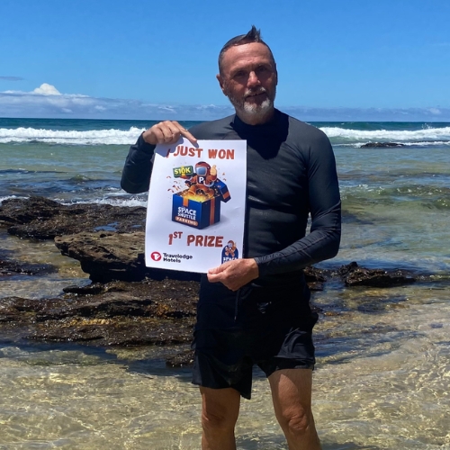 Man in a wetsuit on a rocky beach holds a prize certificate that reads 'Just Won' and '1st Prize' beside a space-themed graphic.