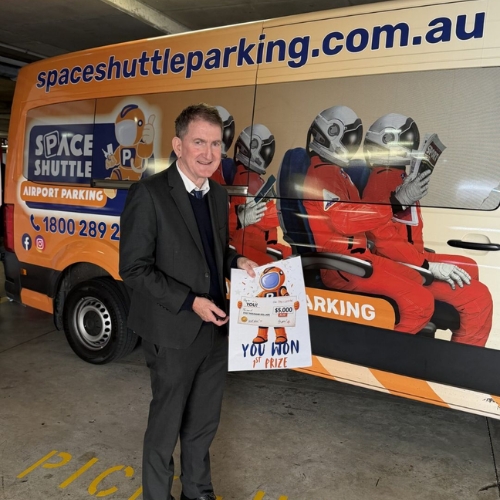 Man in a suit posing beside a bright orange 'Spaceshuttleparking' van, holding a prize certificate that reads 'You won' and '$5,000 prize' (space-themed vehicle).