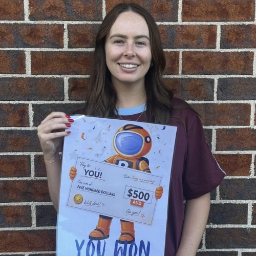 Smiling woman standing against a brick wall, holding a poster featuring a cartoon astronaut and a $500 AUD prize check that says 'You won'.
