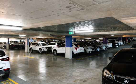 Underground parking garage with rows of parked cars, concrete pillars, and fluorescent lights on a low ceiling.