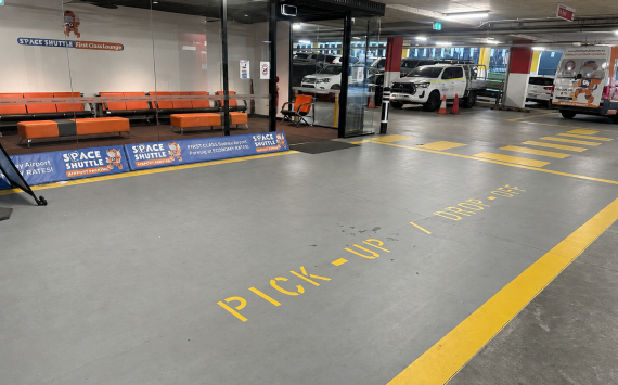 Airport pickup zone with orange seating area behind glass and signs for Space Shuttle airport parking services nearby