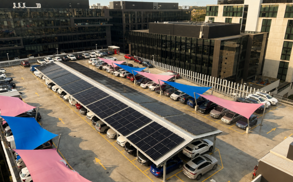 Aerial view of a parking lot with carport solar panels and colorful sail shade canopies beside glass-front office buildings.