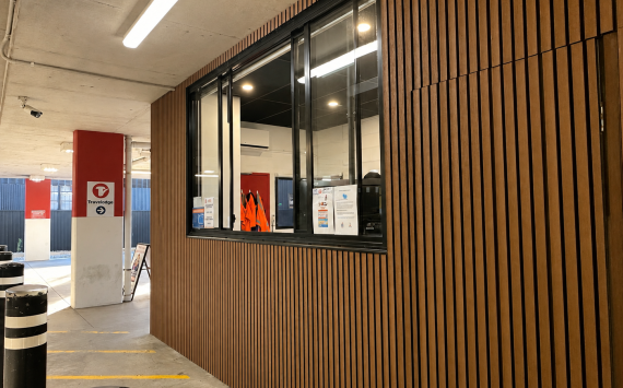 Indoor parking area with a large windowed service desk set into a wood-slatted wall; orange safety vests and posted notices are visible inside the room.