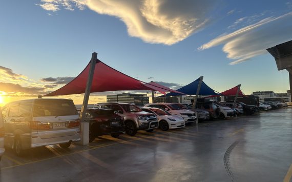 Sunset over a rooftop parking lot with red and blue shade sails and many parked cars.